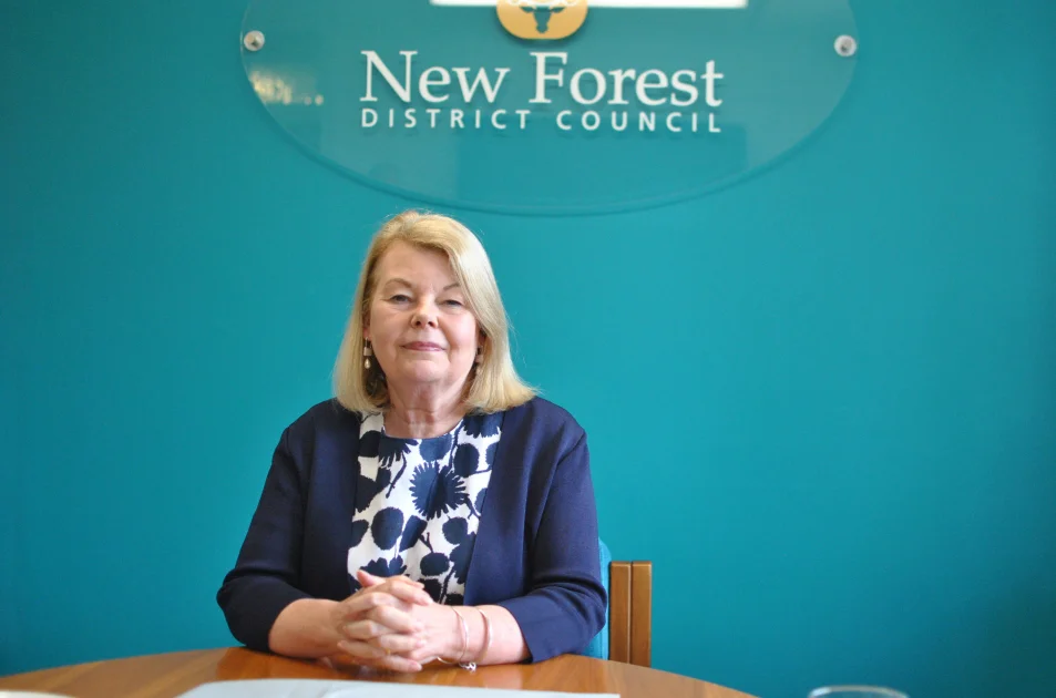 A woman with blonde hair, wearing a blue and white patterned blouse and navy jacket, sits at a table in front of a sign reading New Forest District Council on a teal wall.