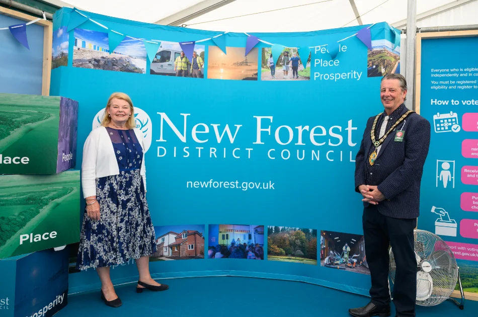 A woman and a man wearing a ceremonial chain stand smiling in front of a New Forest District Council display with photos, the council website, and stacked boxes reading People, Place, and Prosperity.