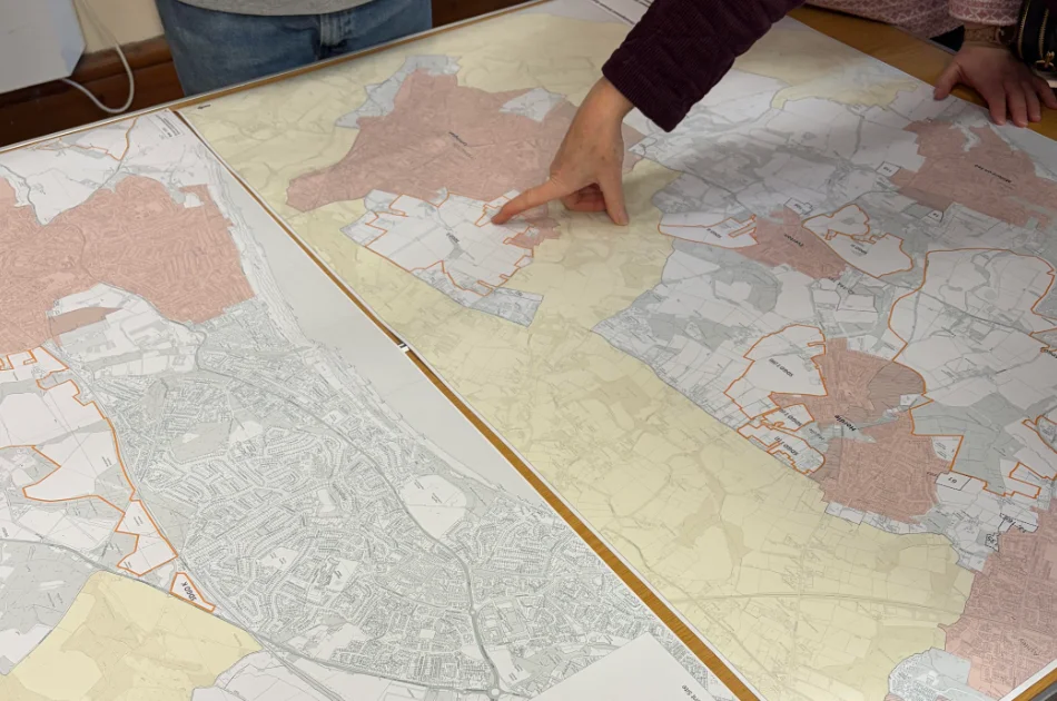 A person points to a specific area on a large map spread out on a table, while another person's hands rest on the map. The map shows regions highlighted in various colours.